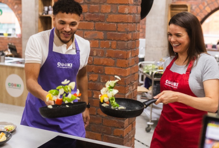 A man and a woman tossing vegetable in a frying pan and enjoying a cookery class.
